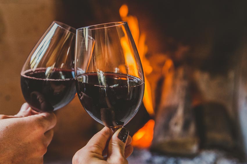 Zwei Hände mit jeweils einem Glas Rotwein stoßen an, im Hintergrund Kaminfeuer