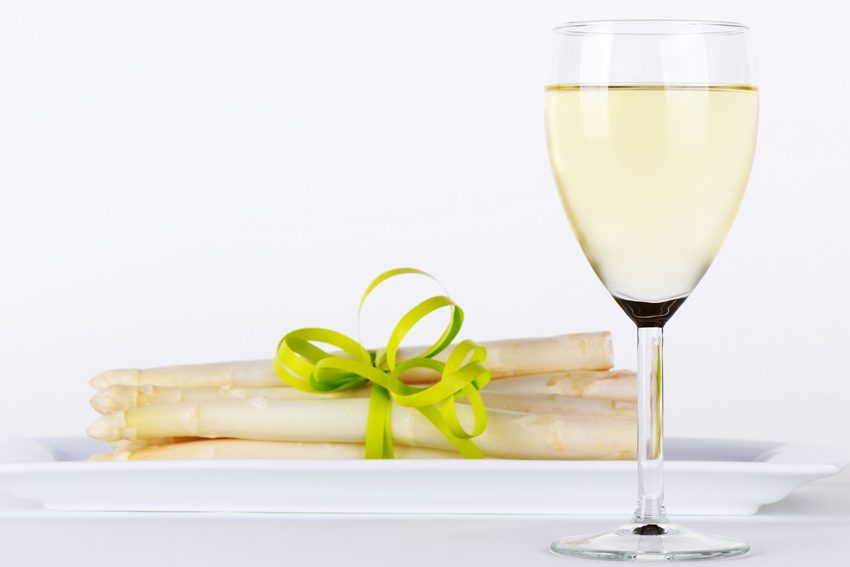 weißer Spargel mit einer grünen Schleife zusammengebunden auf einem Teller, daneben ein Glas Weißwein